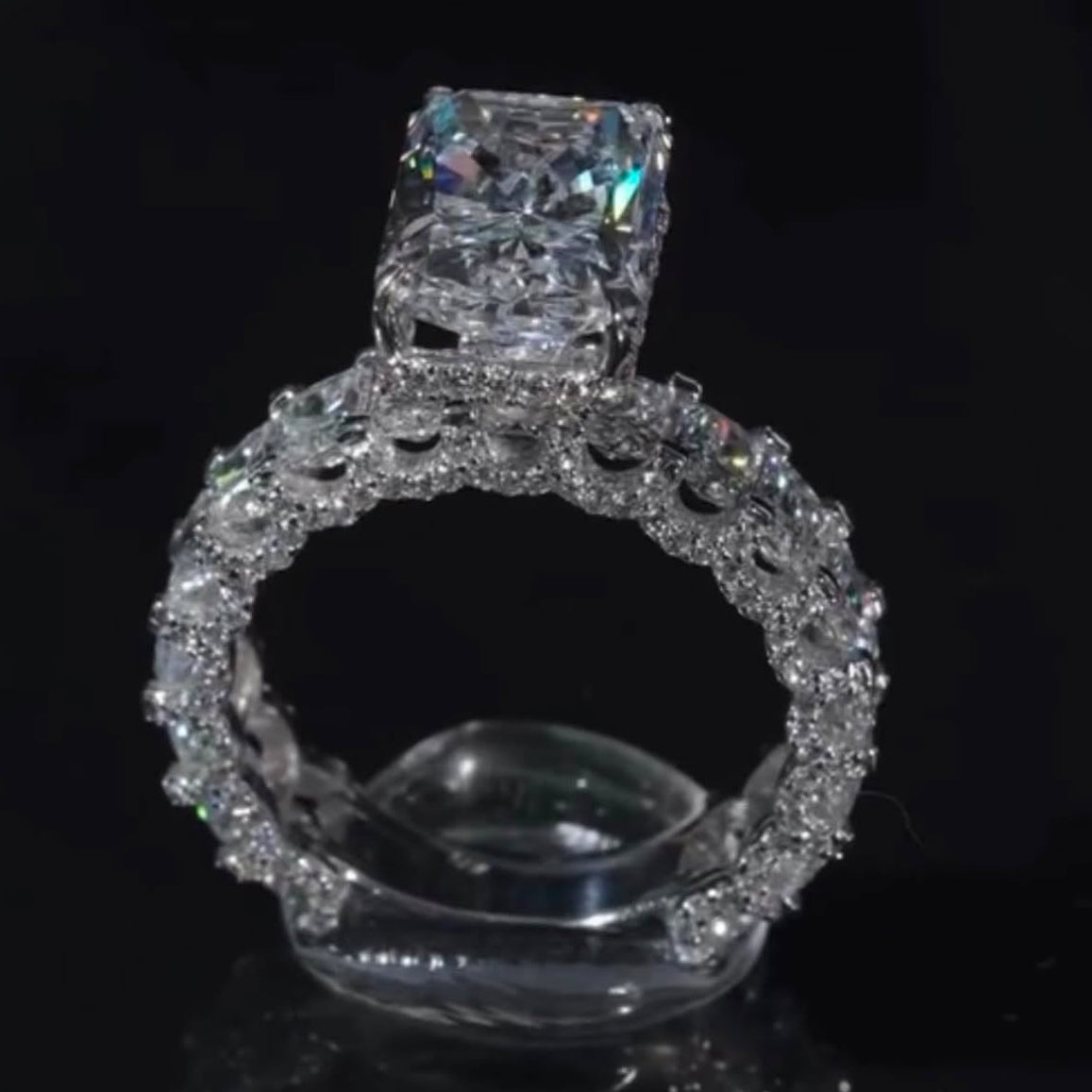 A radiant cut diamond engagement ring with a halo setting, displayed on a clear stand against a black reflective background.