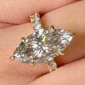 A close-up of a hand wearing a silver ring with a marquise-cut diamond, along with matching diamond bands on other fingers and a diamond bracelet, against a dark background.