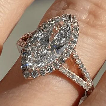 A close-up of a hand wearing a rose gold ring with a large marquise-cut diamond and a halo of smaller diamonds. The ring is shown on the middle finger against a soft, out-of-focus gray background.