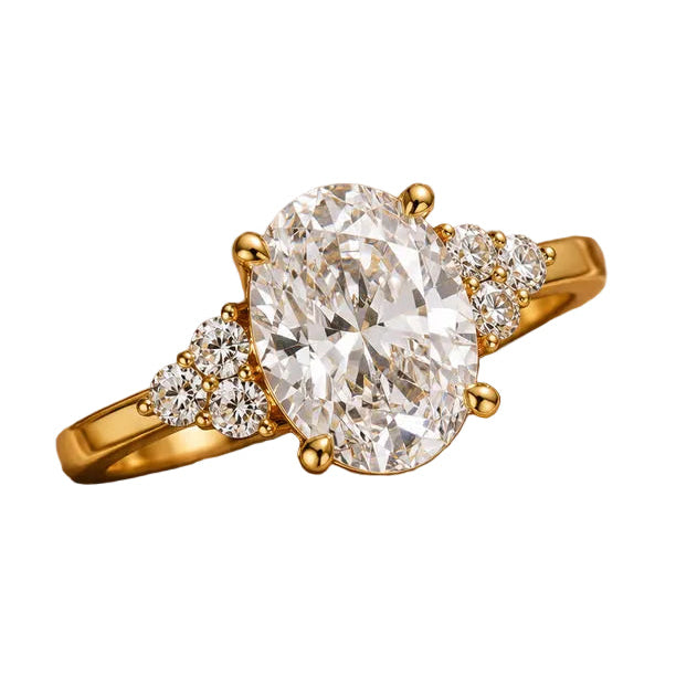 A yellow gold ring featuring a large oval-cut clear gemstone in a prong setting, accented by small round stones on the shoulders, displayed on a textured gold jewelry box with dried flowers.