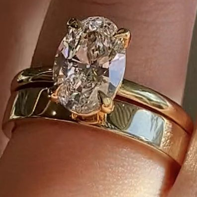 A close-up of a hand wearing a gold ring with a large oval clear gemstone, layered with a slim gold band.