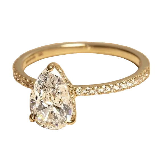 A yellow gold ring featuring a large pear-cut clear gemstone in a prong setting, with small pavé diamonds along the band, displayed on a textured beige surface.