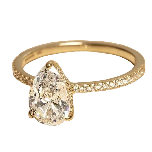 A yellow gold ring featuring a large pear-cut clear gemstone in a prong setting, with small pavé diamonds along the band, displayed on a textured beige surface.