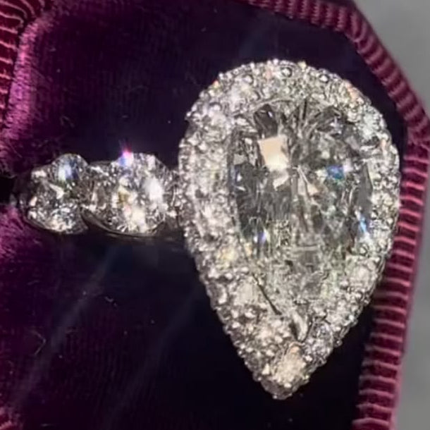 A pear-shaped diamond ring with a halo and a smaller matching diamond on the band, displayed in an open purple velvet ring box held in a hand.